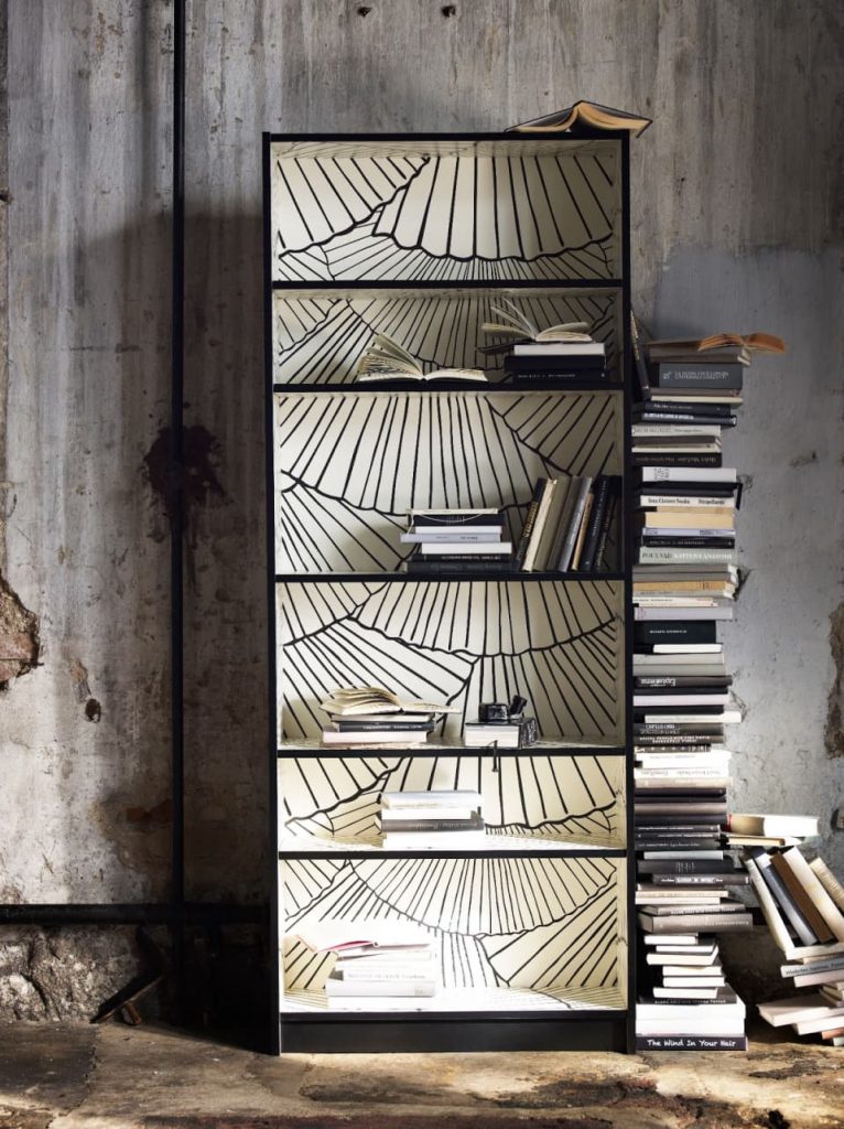Estante antiga com papel de parede aplicado no fundo e livros empilhados ao lado da estante.