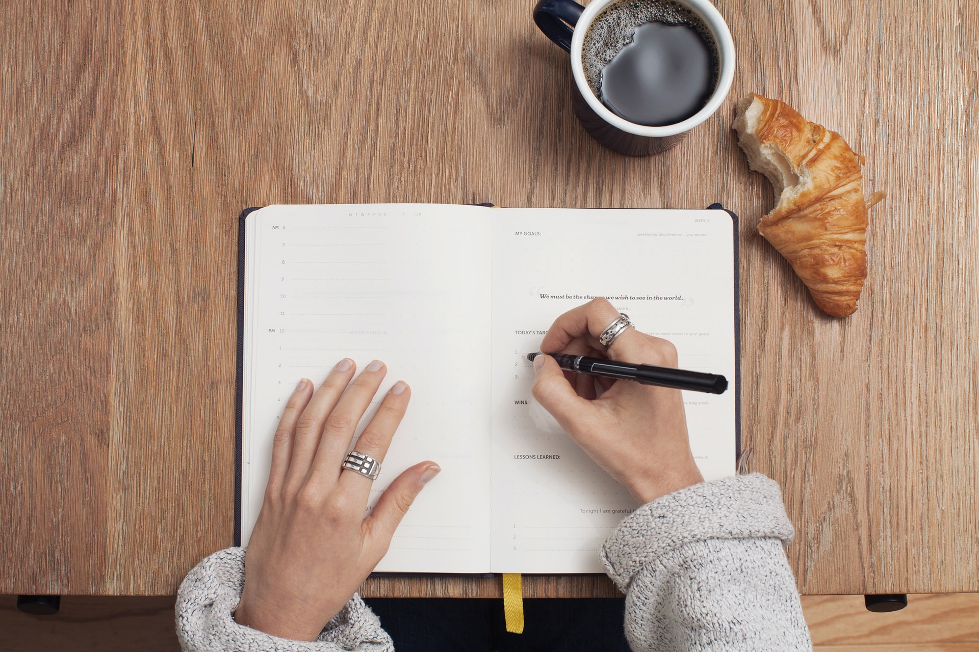 Mulher escrevendo em um caderno, com um café um um croissant sobre a mesa