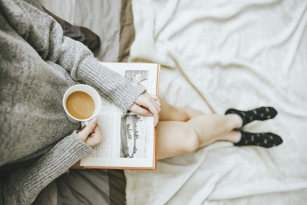 Mulher sentada na cama, com as pernas cruzadas, com um livro aberto e uma xícara de café na mão.