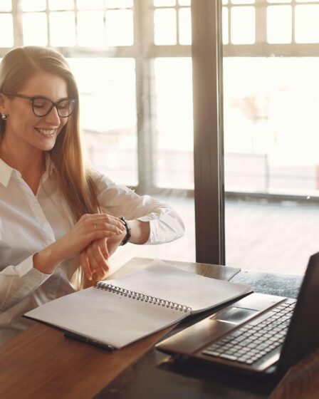 smiling young female entrepreneur checking time on wristwatch while working on laptop in modern workspace