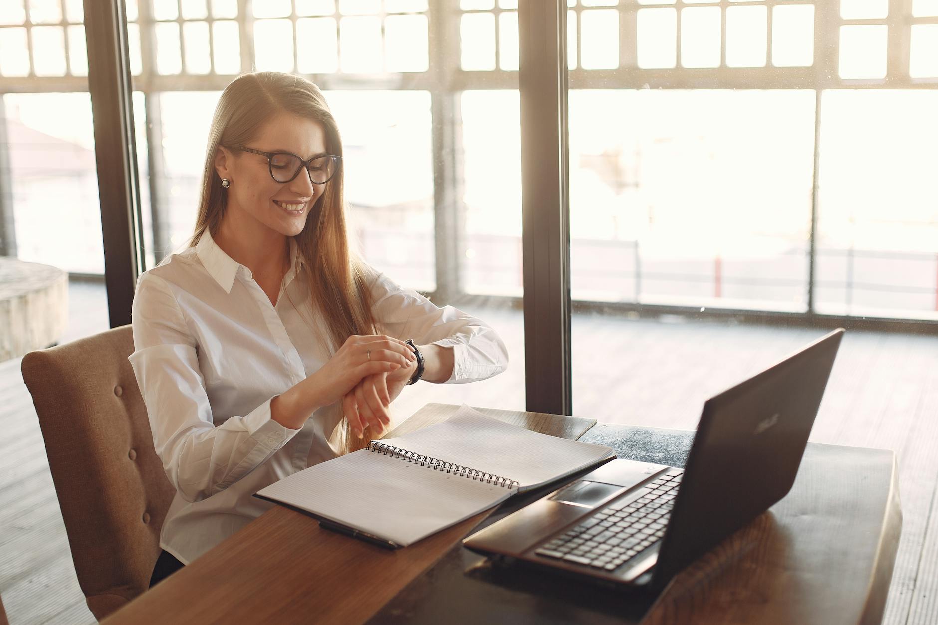 smiling young female entrepreneur checking time on wristwatch while working on laptop in modern workspace